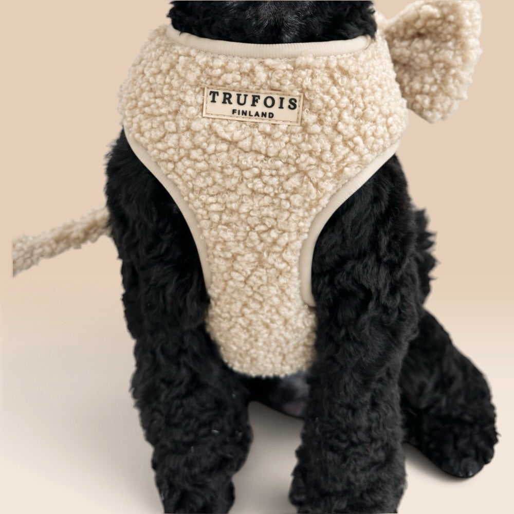 A black curly-haired dog wearing a cream sherpa pet harness with attached ears and a small brand patch reading TRUFOIS FINLAND.