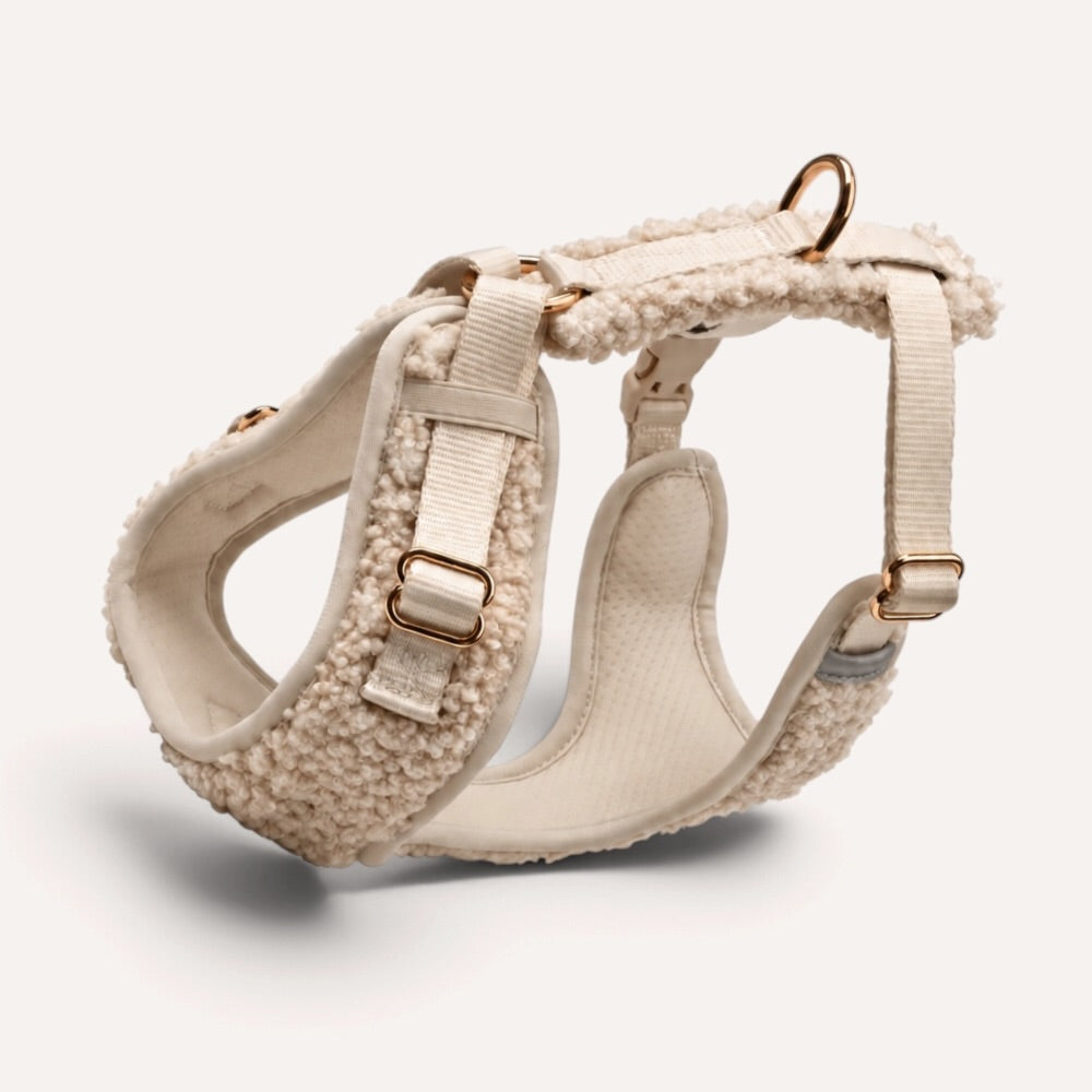 A cream-colored dog harness featuring a plush sherpa fleece texture, adjustable webbing straps, and gold-tone metal hardware, shown on a light beige background.