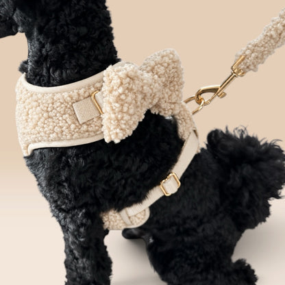 A black curly-haired dog wearing a cream sherpa harness with a matching bow and leash, shown against a solid beige background.