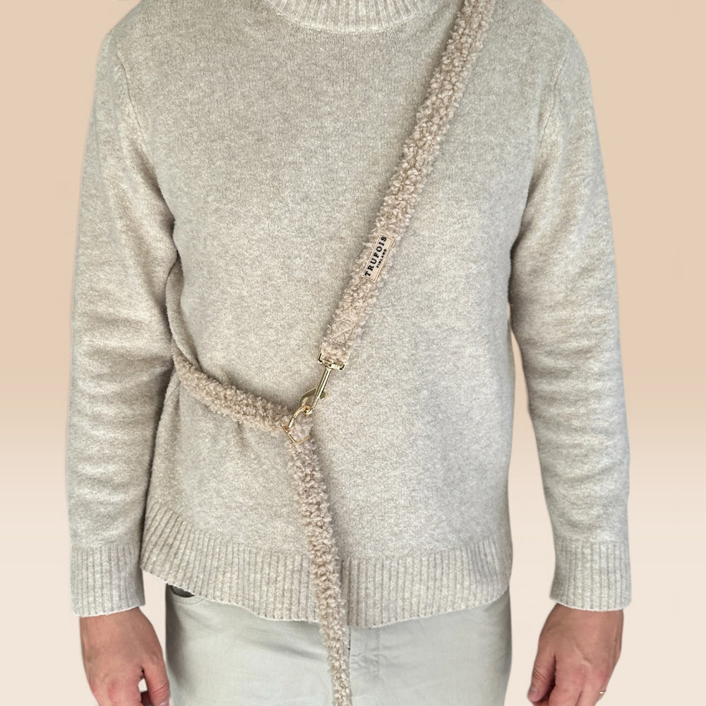 A person wearing a light heather gray knit sweater and pale pants, shown from neck to upper thighs. A chunky knitted beige crossbody dog leash with a small black brand tag and gold-tone hardware crosses the sweater.